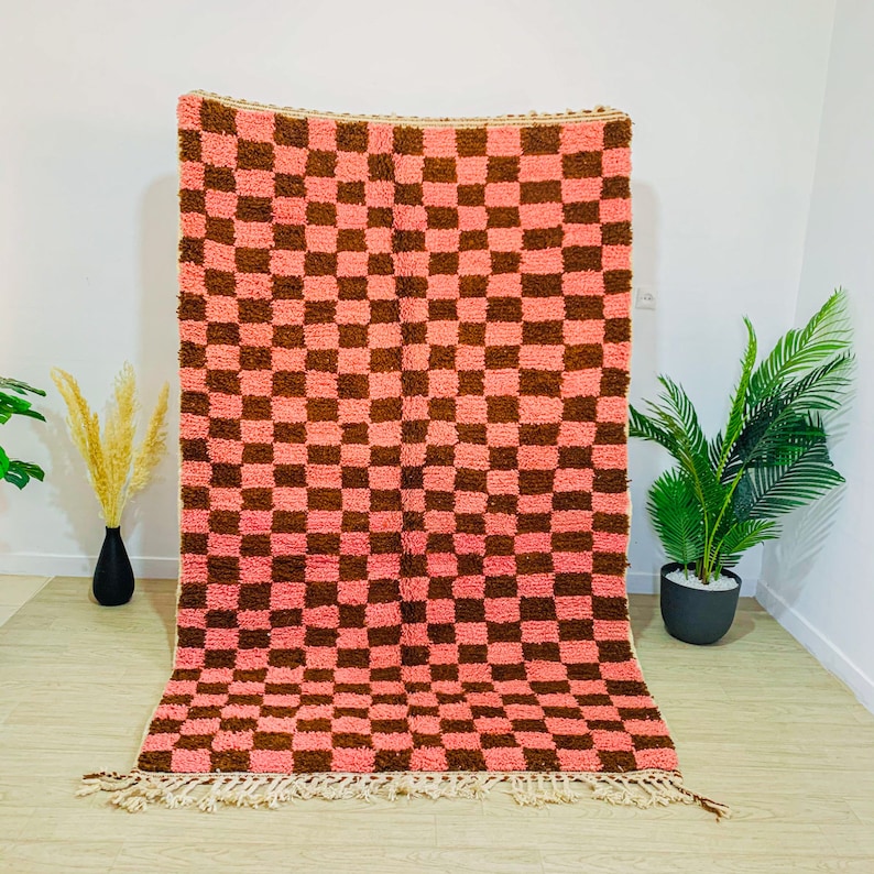May include: A pink and brown checkered rug with a fringe border. The rug is made of a soft, fluffy material.