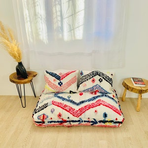 May include: A white, pink and blue patterned floor cushion with two matching pillows. The cushion is made of a soft, fluffy material and has a zig-zag pattern.