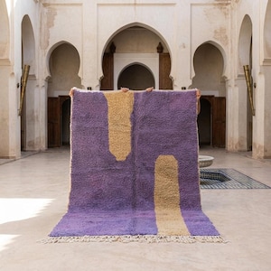 May include: A large, rectangular rug with a plush texture. The rug features a gradient of purple hues and two vertical, abstract shapes in a light tan color. The rug has a fringe along the bottom edge.