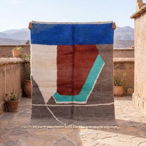 May include: A rectangular rug with a geometric design. The rug features a blue top section, a central maroon shape, and a teal and white accent. The bottom is gray with a white line design. The rug is held up against a backdrop of mountains and a clear sky.
