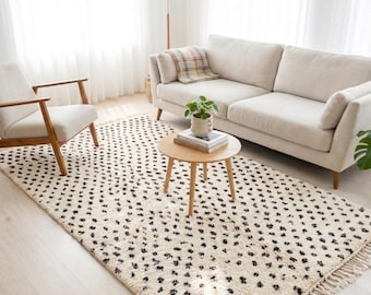 May include: A living room scene featuring a cream-colored sofa, armchair, and a round wooden coffee table. A patterned rug with black spots covers the floor. A plaid throw blanket rests on the sofa, and a potted plant sits on the table.