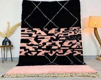 Könnte beinhalten: Ein großer, rechteckiger Teppich mit schwarzem Hintergrund und einem geometrischen Design in Weiß und Rosa. Der Teppich hat einen rosafarbenen Rand mit Fransen. Der Teppich besteht aus einem dicken, strukturierten Material.