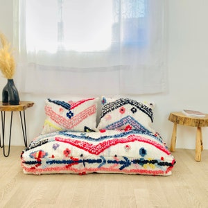 May include: A white, pink and blue patterned floor cushion with two matching pillows. The cushion is made of a soft, fluffy material and has a geometric design.