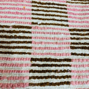 May include: Close-up of a woven rug with a pattern of brown and pink stripes. The rug is made of a soft, textured material.