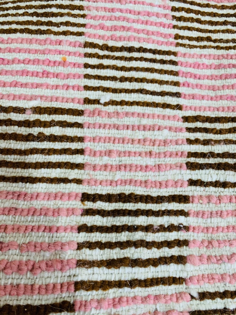 May include: Close-up of a woven rug with a pattern of brown and pink stripes. The rug is made of a soft, textured material.
