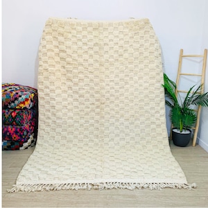 May include: A white woven rug with a checkered pattern and fringe. The rug is laid out on a wooden floor.