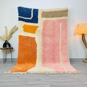 May include: A large, rectangular rug with an abstract design. The rug features a cream-colored background with patches of orange, pink, blue, and tan. The rug has a shaggy texture and a fringe along the bottom edge.