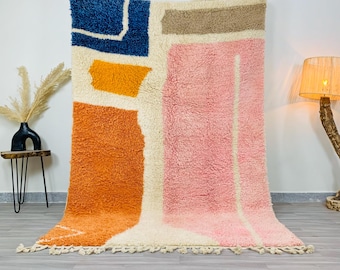 Puede incluir: Una alfombra rectangular grande con un diseño abstracto. La alfombra presenta un fondo color crema con parches de color naranja, rosa, azul y beige. La alfombra tiene una textura peluda y una franja en el borde inferior.