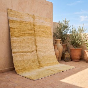Könnte beinhalten: Ein großer, rechteckiger Teppich in Gelb- und Cremetönen mit strukturierter Oberfläche. Der Teppich lehnt an einer hellbraunen Wand. Terrakottatöpfe und Pflanzen sind im Hintergrund zu sehen. Der Teppich hat ein Streifenmuster.