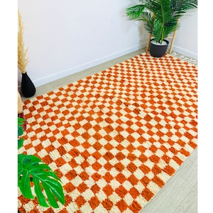 May include: A large, orange and white checkered rug with a fluffy texture. The rug is made of wool and has a fringe border.