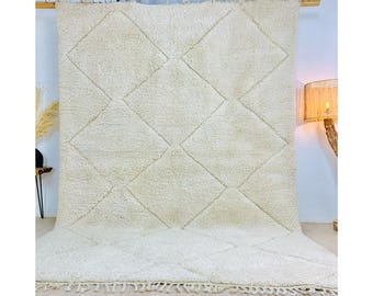 May include: A white, hand-woven Moroccan rug with a diamond pattern. The rug has a fringe border and is made of natural fibers.