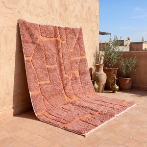 May include: A large, rectangular, coral-colored rug with a geometric design. The rug features raised, textured patterns in a lighter orange hue, creating a striking contrast. The rug is leaning against a wall, showcasing its size and design.