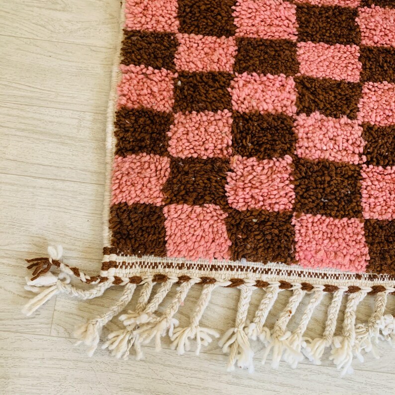 May include: A pink and brown checkered rug with a fringe border. The rug is made of a soft, fluffy material.