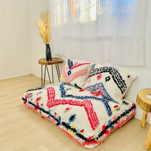 May include: A white floor cushion with a colourful geometric pattern. The cushion is made of a soft, fluffy material and has a fringe border. Two colourful pillows are placed on top of the cushion.