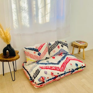 May include: A white, pink and blue patterned pouffe with two matching cushions. The pouffe is made of a soft, fluffy material and has a geometric design.