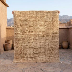 May include: A large, rectangular beige rug with a textured, grid-like pattern. The rug is held up against a backdrop of a stone building and mountains. The rug's thick pile suggests a soft, comfortable texture, suitable for a living room or bedroom.