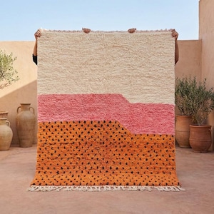 May include: A rectangular rug with a cream-colored top section, a pink middle section, and an orange bottom section with black dots. The rug has a textured, shaggy appearance and a fringe along the bottom edge. The rug is held up by unseen hands.