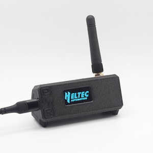 Puede incluir: Un dispositivo electrónico negro con una pequeña pantalla que muestra el texto "HELTEC AUTOMATION" en azul. Tiene una antena negra y un cable negro enchufado. El dispositivo está sobre un fondo blanco.