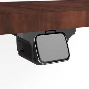 May include: A black under-desk mount with a gray speaker attached to it. The mount is attached to a wooden surface.