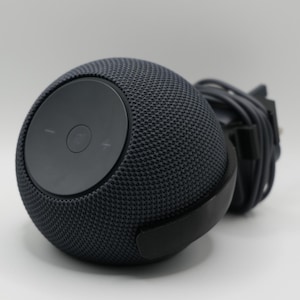 Puede incluir: Un altavoz inteligente esférico gris oscuro con un exterior de tela de malla. La parte superior presenta un panel de control circular con botones de volumen. Un cable de alimentación negro está conectado.