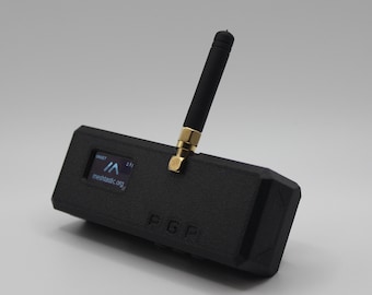 May include: A black, rectangular device with a small screen displaying the text "meshtactic.org" and a small antenna. The device has a gold-colored connector and the text "F.G.F" printed on the side.
