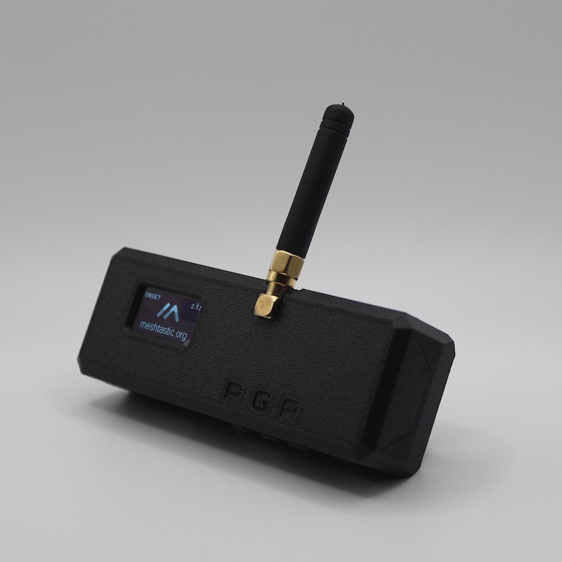 May include: A black, rectangular device with a small screen displaying the text "meshtactic.org" and a small antenna. The device has a gold-colored connector and the text "F.G.F" printed on the side.