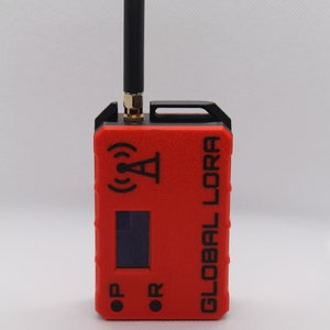 May include: A red and black handheld device with a black antenna, a small screen, and the text "GLOBAL LORA" printed on the side. The device has two buttons labeled "OP" and "OR".