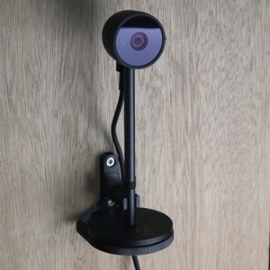 May include: A black webcam mounted on a wooden surface. The webcam has a circular lens and is connected to a stand and base. A cable extends from the base. The webcam is designed for video conferencing and online meetings.