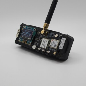 May include: A black circuit board with a small screen displaying text, a gold antenna, and the text "LILYGO&reg; Model: LORA32 868/915MHz" printed on the board. The text on the screen reads "MESNTASTIC Mar030 Set the region using the Menu/Android app. Flash or CU deat"