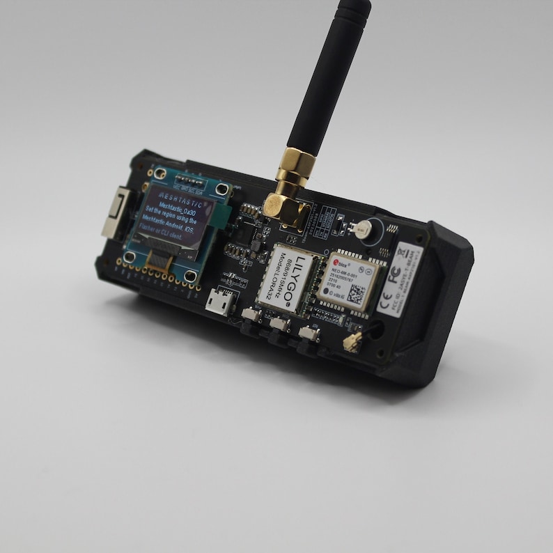 May include: A black circuit board with a small screen displaying text, a gold antenna, and the text "LILYGO&reg; Model: LORA32 868/915MHz" printed on the board. The text on the screen reads "MESNTASTIC Mar030 Set the region using the Menu/Android app. Flash or CU deat"