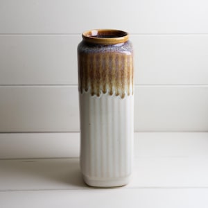 May include: A cylindrical ceramic vase featuring a vertical ribbed texture on the lower portion. The upper section is finished with a reactive glaze in shades of brown and purple that drips down over the off-white base, creating a distinct contrast.