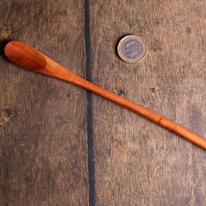 May include: A long, slender wooden spoon with a rounded head, displayed on a wooden surface. The spoon is a warm reddish-brown colour. A coin is also present, providing a size reference. The spoon appears to be handmade.