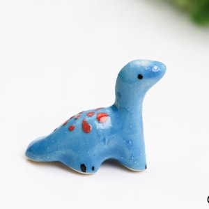 Könnte beinhalten: Kleine, hellblaue Keramik-Dinosaurierfigur mit roten Flecken auf dem Rücken. Der Dinosaurier hat einen langen Hals und kleine schwarze Augen und Füße. Dieser dekorative Artikel ist eine charmante Ergänzung für jede Sammlung.