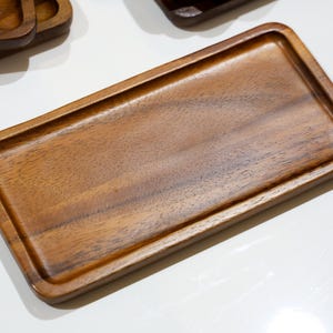 Op de afbeelding: Een rechthoekige houten dienblad met een opstaande rand. De lade is gemaakt van donkerbruin hout met een zichtbare nerf. Verschillende soortgelijke laden zijn gedeeltelijk zichtbaar op de achtergrond. De lade is ongeveer 30 cm lang en 15 cm breed.