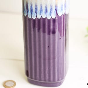 May include: A tall, cylindrical vase with a ribbed, deep purple body. The top features a scalloped design in shades of blue, white, and gold. The vase is likely ceramic and suitable for home decor.