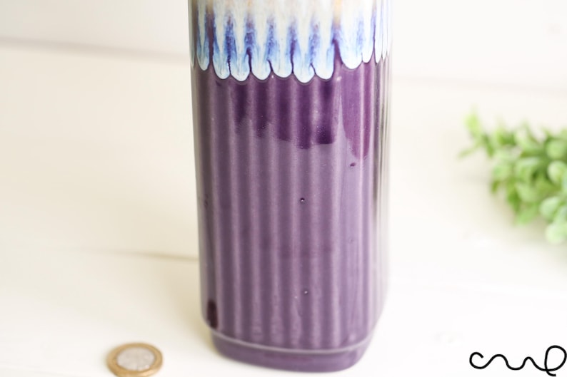 May include: A tall, cylindrical vase with a ribbed, deep purple body. The top features a scalloped design in shades of blue, white, and gold. The vase is likely ceramic and suitable for home decor.