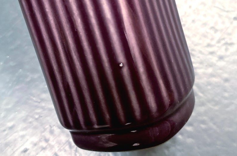 May include: Close-up of a dark purple ceramic container with vertical ribbed detailing. The container has a rounded base and a slightly flared top. The surface is glossy, reflecting light.