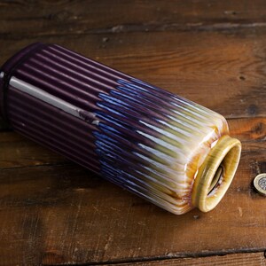 May include: A cylindrical ceramic vase with a ribbed design. The vase transitions from a deep purple to blue and white, with a yellow rim. A coin rests on the wooden surface next to the vase. Decorative home decor.