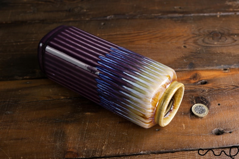 May include: A cylindrical ceramic vase with a ribbed design. The vase transitions from a deep purple to blue and white, with a yellow rim. A coin rests on the wooden surface next to the vase. Decorative home decor.