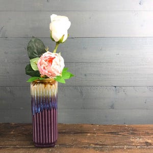 Op de afbeelding: Een decoratieve vaas met een verticaal geribbeld ontwerp in tinten paars, blauw en bruin. De vaas bevat een roze pioenroos en een witte roos. De opstelling staat tegen een grijze houten achtergrond.