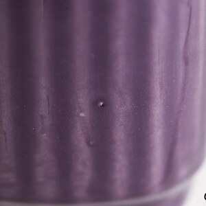 May include: Close-up of a purple ceramic container with vertical ribbed detailing. The container has a cylindrical shape and a smooth, glossy finish. A small, dark spot is visible on the surface.