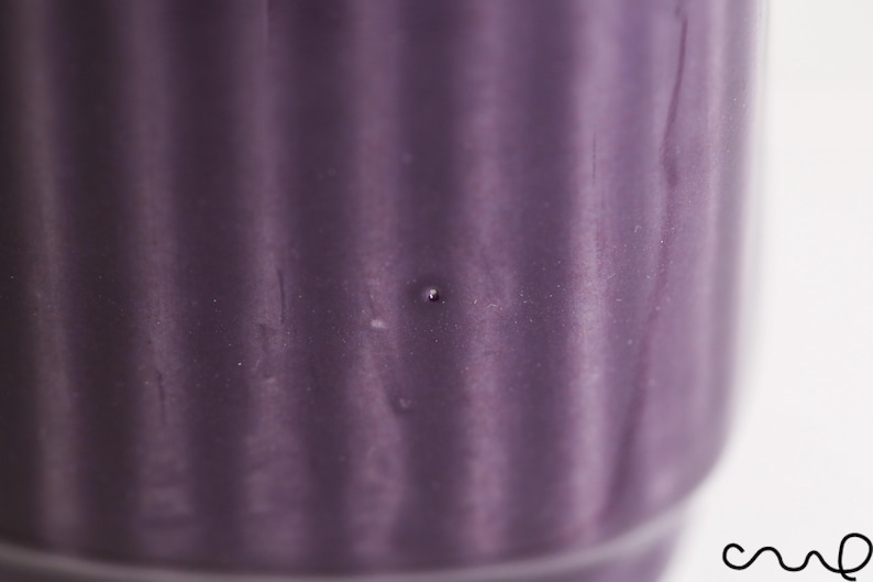 May include: Close-up of a purple ceramic container with vertical ribbed detailing. The container has a cylindrical shape and a smooth, glossy finish. A small, dark spot is visible on the surface.