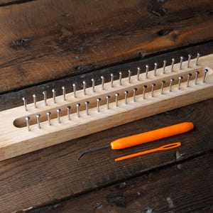 Peut inclure: Un métier à tricoter rectangulaire en bois clair doté de deux rangées parallèles de tiges métalliques régulièrement espacées. Le métier est accompagné de deux outils en plastique orange : un crochet à pointe métallique et une aiguille.
