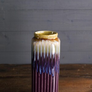 May include: A tall, cylindrical ceramic vase with a ribbed texture. The vase features a color gradient, transitioning from a yellow rim to white, blue, and a deep purple base. The vase is displayed on a wooden surface.