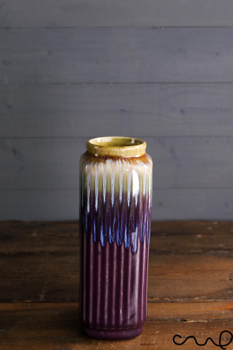 May include: A tall, cylindrical ceramic vase with a ribbed texture. The vase features a color gradient, transitioning from a yellow rim to white, blue, and a deep purple base. The vase is displayed on a wooden surface.