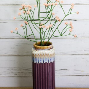 May include: A tall, ribbed, plum-colored ceramic vase with a cream, blue, and brown rim holds a bundle of artificial pink flowers with green stems. The vase sits on a wooden surface against a white-painted wood background.