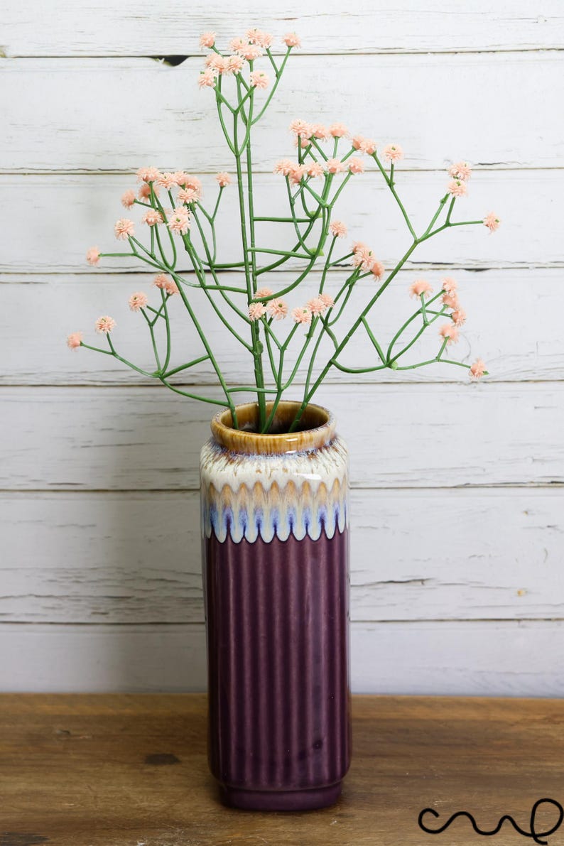 May include: A tall, ribbed, plum-colored ceramic vase with a cream, blue, and brown rim holds a bundle of artificial pink flowers with green stems. The vase sits on a wooden surface against a white-painted wood background.