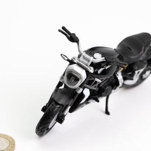 Peut inclure: Modèle de moto noire avec des détails argentés, notamment le guidon, le phare et l'échappement. La moto miniature a un design élégant et est posée sur un fond blanc. Une pièce de monnaie est au premier plan.