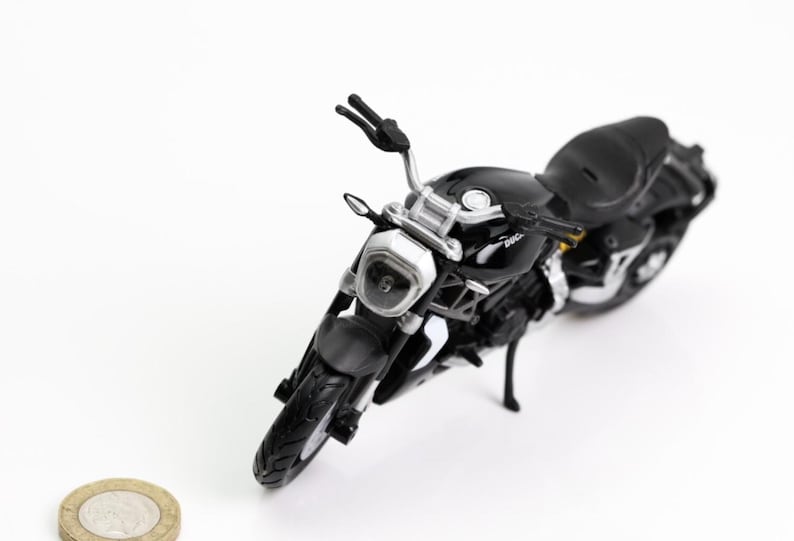 May include: A black motorcycle model with silver accents, including the handlebars, headlight, and exhaust. The miniature bike has a sleek design and is set against a white background. A coin is in the foreground.