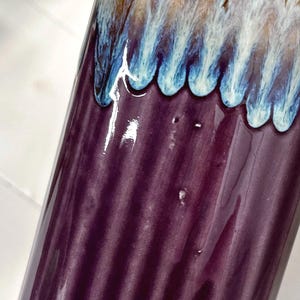 May include: A cylindrical ceramic vase with a glossy finish. The vase is primarily a deep purple color with vertical ridges. The top edge features a decorative band of blue, white, and brown glaze.